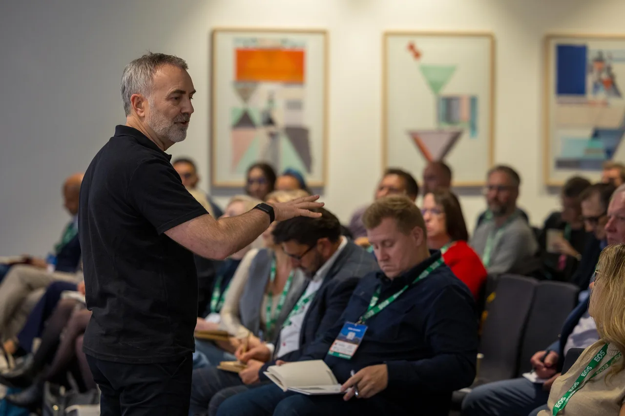A facilitator standing mid-sentence in front of a seated group at an EOS session, notebooks open across the room.