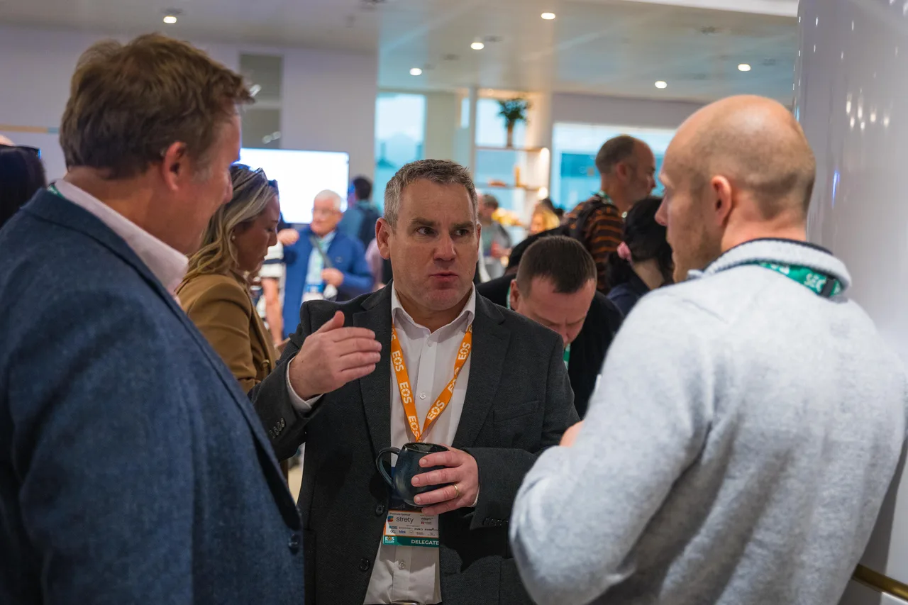 Four people standing in a close huddle at an EOS conference, one mid-explanation with a coffee in hand while the others listen.